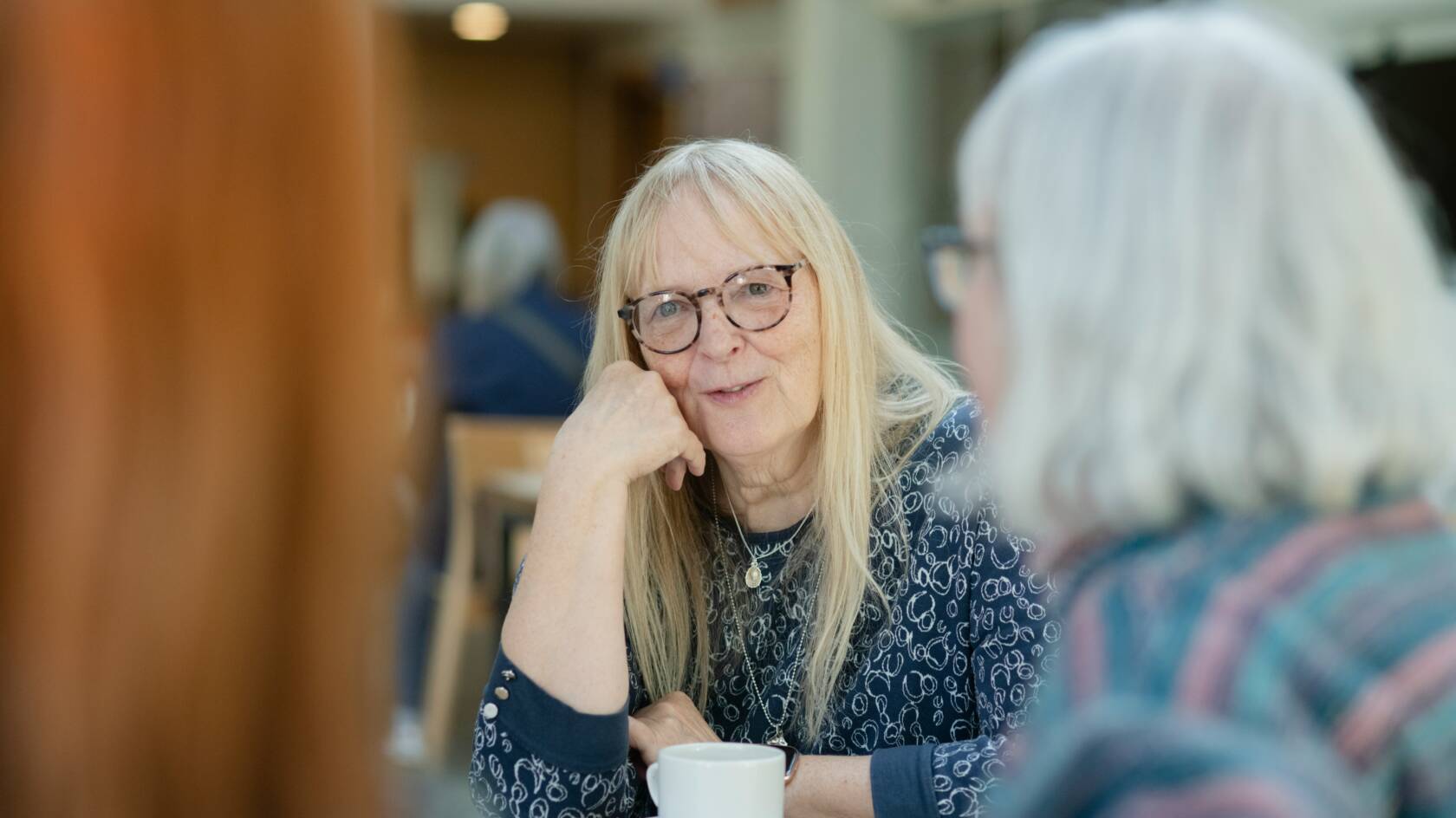 Iäkäs nainen nojaa kämmeneensä poskellaan ja puhuu muille naisille. Edessään hänellä on kahvikuppi. Naisella on pitkät blondit hiukset ja silmälasit.
