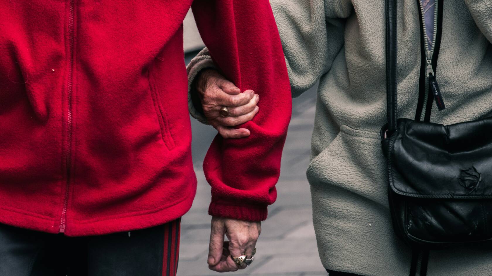 Lähikuva fleeceihin pukeutuneista ikäihmisistä, joilla on ryppyiset kädet. Toinen pitää toista käsipuolesta.