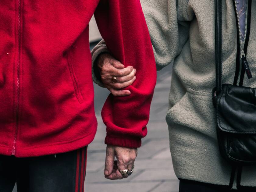 Lähikuva fleeceihin pukeutuneista ikäihmisistä, joilla on ryppyiset kädet. Toinen pitää toista käsipuolesta.