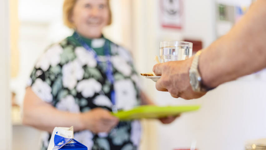 Kuvassa polkkatukkainen, virkapaitaan puettu diakoniatyöntekijä kantaa ruokatarjotinta ja jututtaa samalla toista ihmistä.