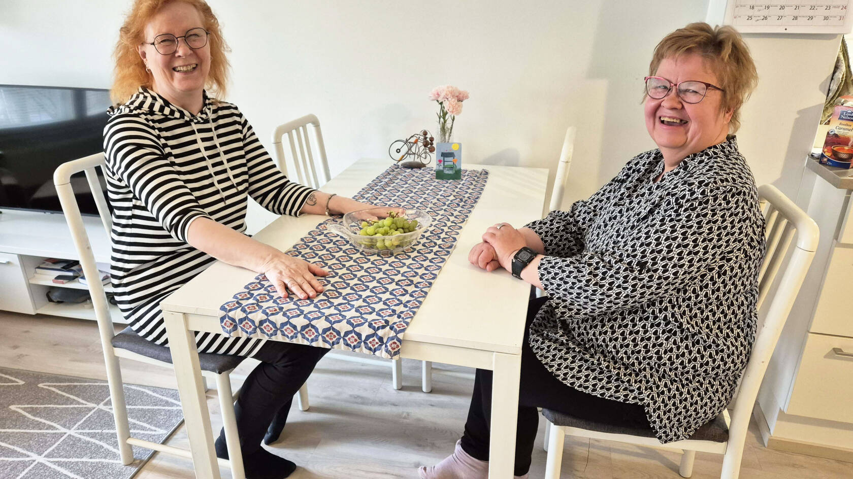 Leila Vilenius ja Anu Kainulainen istuvat keittiön pöydän ääressä toisiaan vastapäätä ja katsovat kameraan hymyillen.
