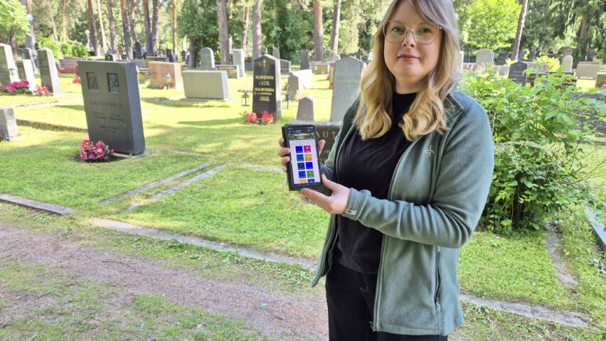 Sarianna Koukkula seisoo hautausmaalla kädessään puhelin, jossa on avoinna hautakarttapalvelun sivu.