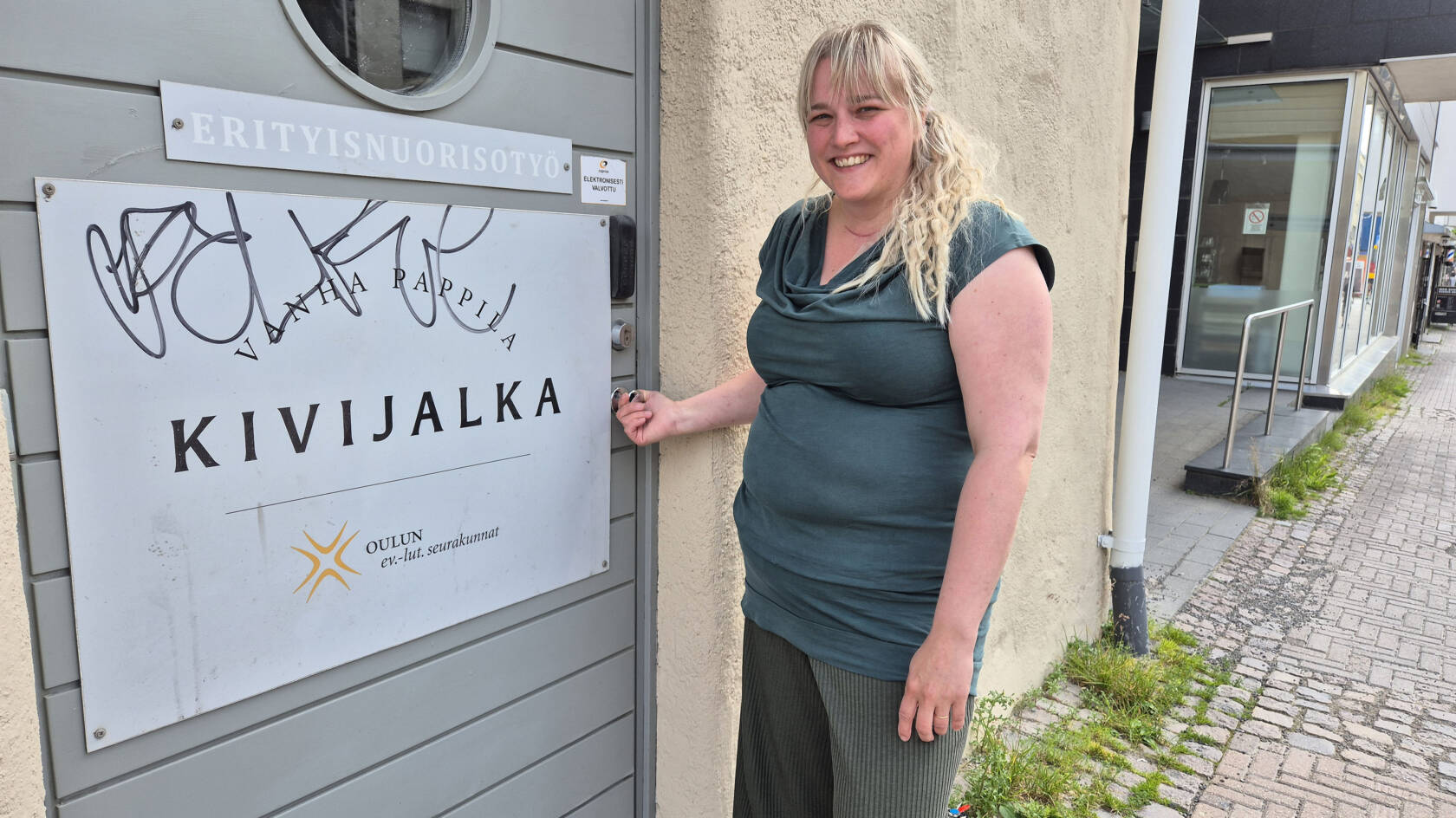 Minna Lappalainen seisoo Kivijalka-tilan oven edessä ja pitää kiinni ovenrivasta.