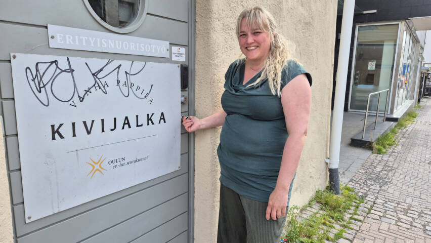 Minna Lappalainen seisoo Kivijalka-tilan oven edessä ja pitää kiinni ovenrivasta.