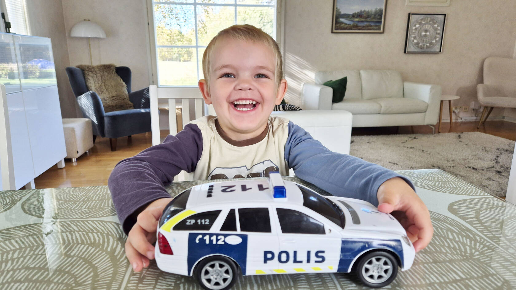 Poika istuu pöydän ääressä, pitää kiinni leluautosta ja hymyilee.
