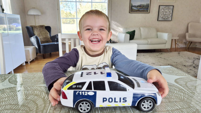 Poika istuu pöydän ääressä, pitää kiinni leluautosta ja hymyilee.