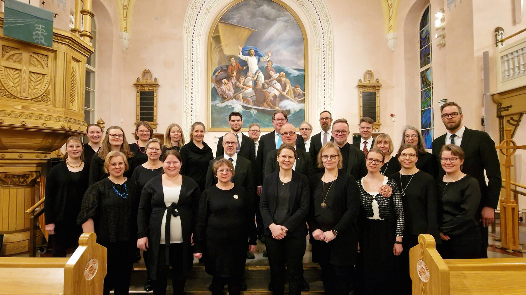 Tummin vaatteisiin pukeutuneet kuorolaiset seisovat asetelmassa alttarin edessä.